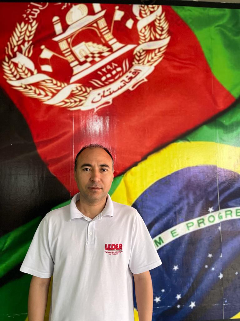 Um homem vestindo camiseta branca com bordado vermelho no canto direito está parado, olhando para a foto. Atrás dele há uma quadro com a bandeira do Brasil e a bandeira do Afeganistão se encostando.